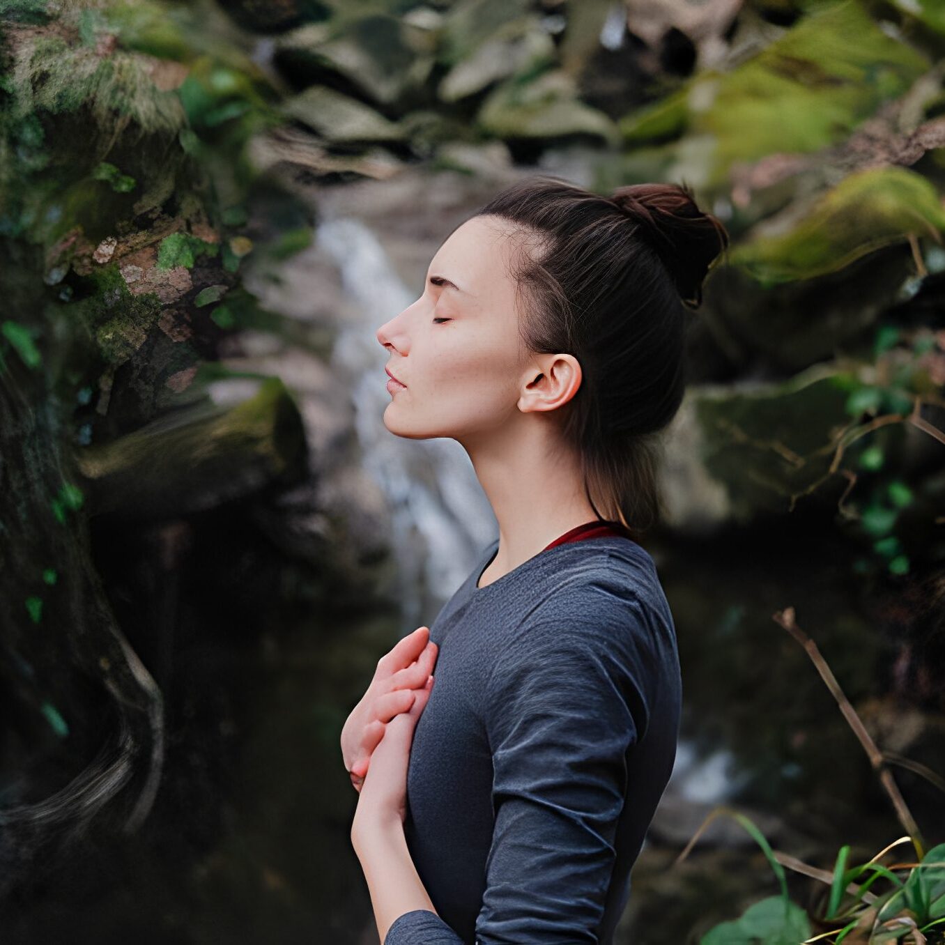 A woman standing peacefully in nature with closed eyes and hand on chest.