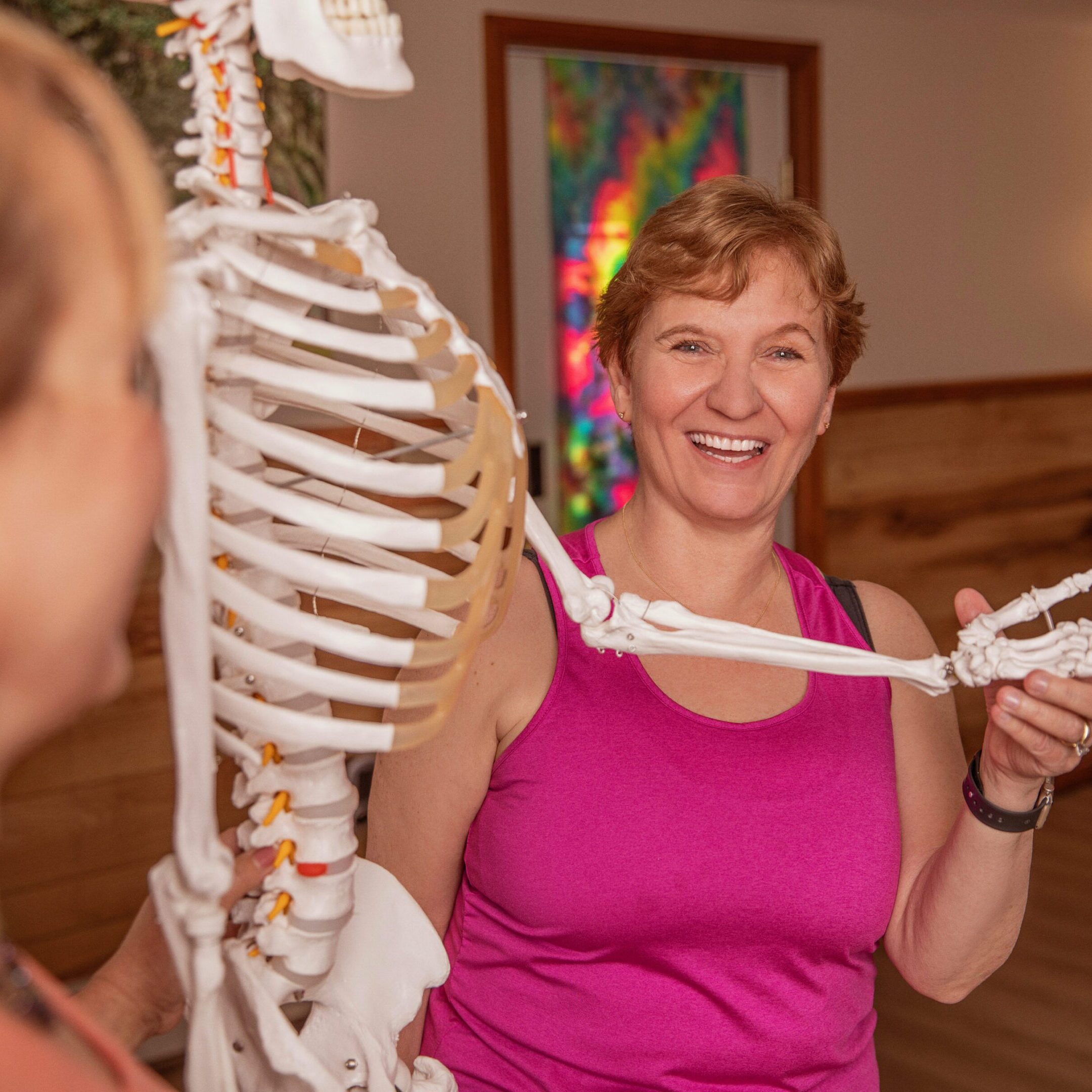A woman smiling while pointing at a skeleton model in a classroom.